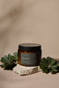Brown Bendalong candle jar with black lid on a stone surface with green leaves, against a beige background