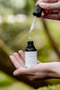 Hand holding a dropper over a bottle labelled Delicious Vibrant Beauty with a blurred green background
