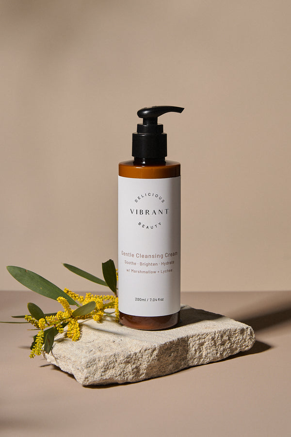 Bottle of 'Delicious Vibrant Beauty' cleanser on a stone surface with yellow flowers on a beige background