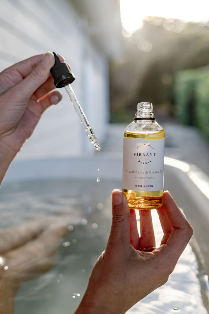 Person holding a bottle of 'Delicious Vibrant Beauty' bath oil with a dropper over water