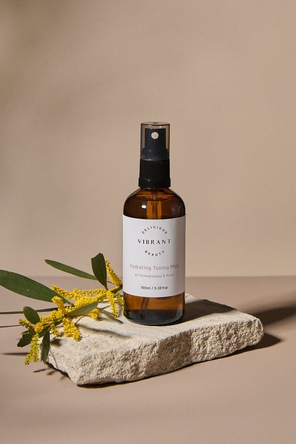 Brown spray bottle labelled Hydrating Toning Mist on a stone surface with yellow flowers on a beige background