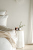 Neatly made bed with a side table featuring a candle and plant in a minimalistic bedroom.