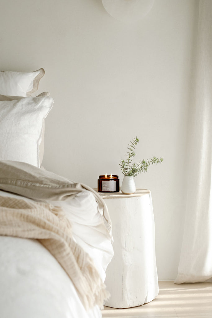 Neatly made bed with a side table featuring a candle and plant in a minimalistic bedroom.