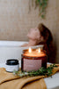 Candle with 'Mollymook' branding on a towel next to a bathtub with a person in the background.