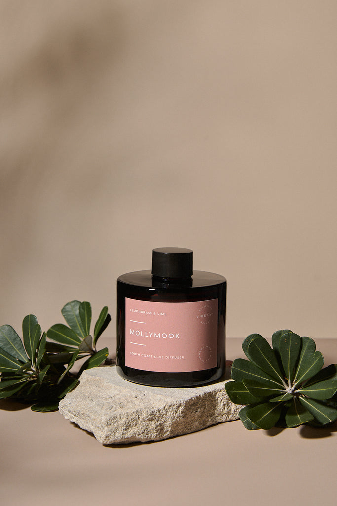 Reed diffuser jar with pink label on a stone surface with green leaves, against a beige background