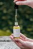 Hand holding a bottle of 'Delicious Vibrant Beauty' product with a dropper against a natural background