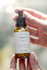 Hand holding a bottle of 'Delicious Vibrant Beauty' elixir with a blurred natural background