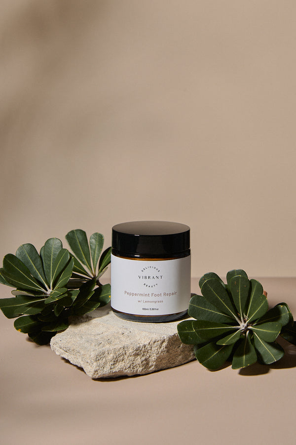 Jar of Peppermint Foot Repair cream on a stone surface with green leaves on a beige background