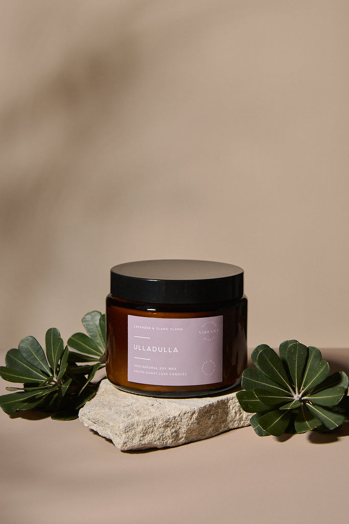 Amber glass candle jar with a mauve label on a stone surface with green leaves, against a beige background