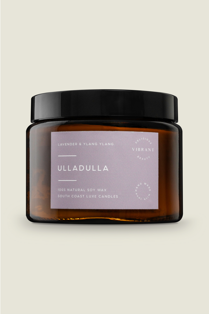 A brown glass jar with a black lid, labeled 'Ulladulla' containing a candle. The label has white and purple text and a circular logo, indicating the scent is Lavender & Ylang Ylang.